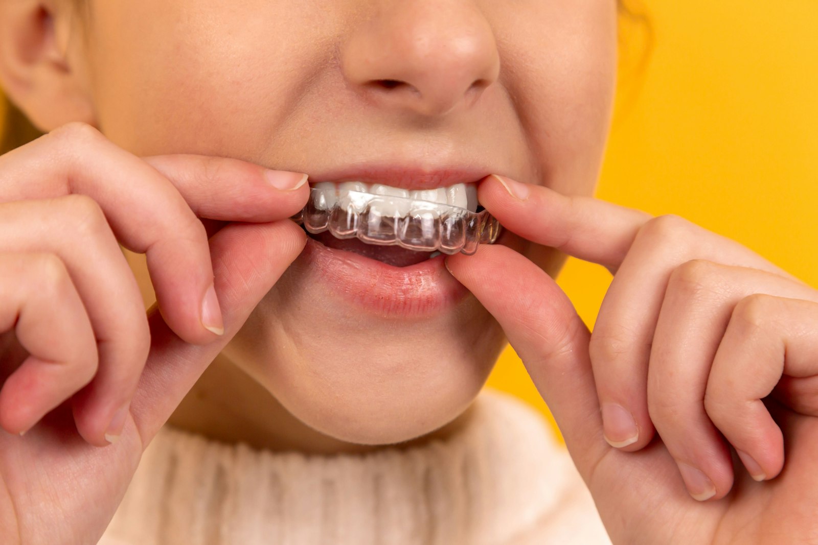 freestocks-Qx5QNarbrUM-unsplash A person inserting a clear plastic dental aligner over their teeth against a yellow background